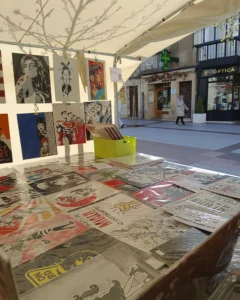 Puesto de feria con cuadros frikis originales expuestos bajo carpa, mesa llena de láminas ilustradas y obras colgadas en pared en mercado artesanal.