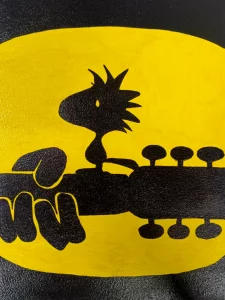 Cuadro de Woodstock tocando una guitarra, representado en silueta negra sobre fondo amarillo y negro de alto contraste.